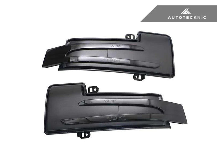 High-resolution detail of AutoTecknic smoked lens housing - aerospace-grade SMD LEDs with amber illumination for Mercedes G-Class on a white background with autotecknic logo on the top right